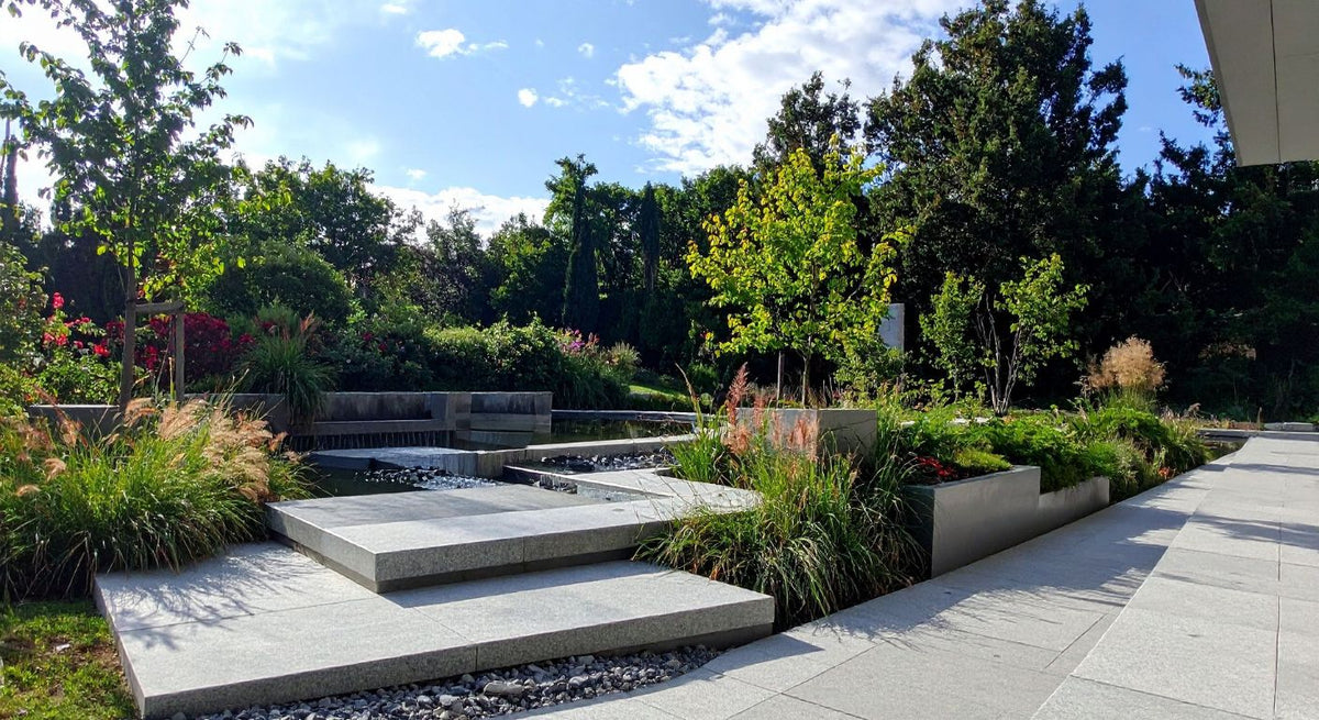 Contemporary garden with clean stone paving, raised planting beds, ornamental grasses, and a simple water feature set among trees.