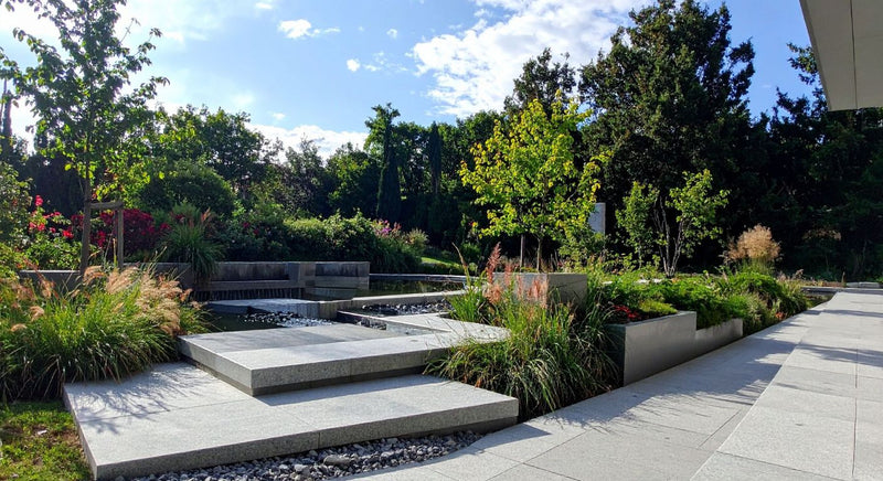 Contemporary garden with clean stone paving, raised planting beds, ornamental grasses, and a simple water feature set among trees.